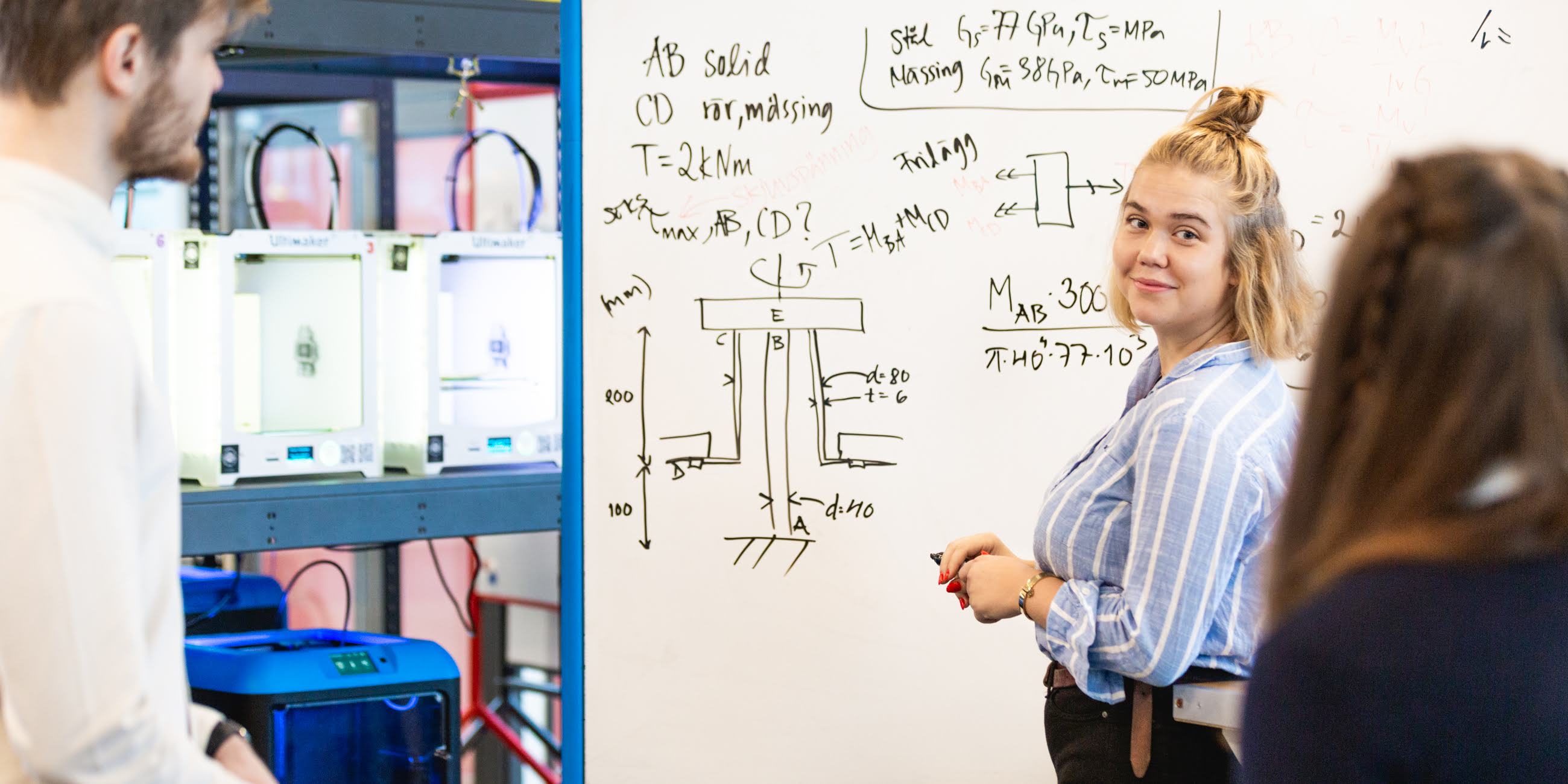 Fotografi på tjej vid whiteboard som kollar tillbaka på två andra studenter