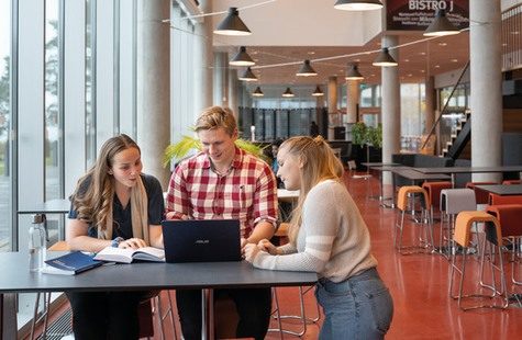 Tre studenter diskuterar vid bord med laptop