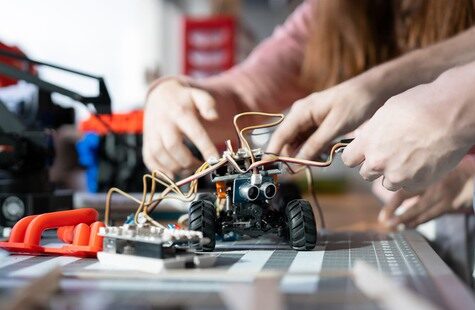 Närbild på två studenter som håller på att bygga en liten robot med hjul och kameror till ögon