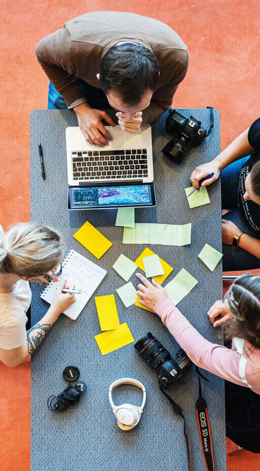 Foto ovanifrån på ett bord där fyra studenter sitter och studerar. På bordet ligger en kameror, postit-lappar och en laptop.